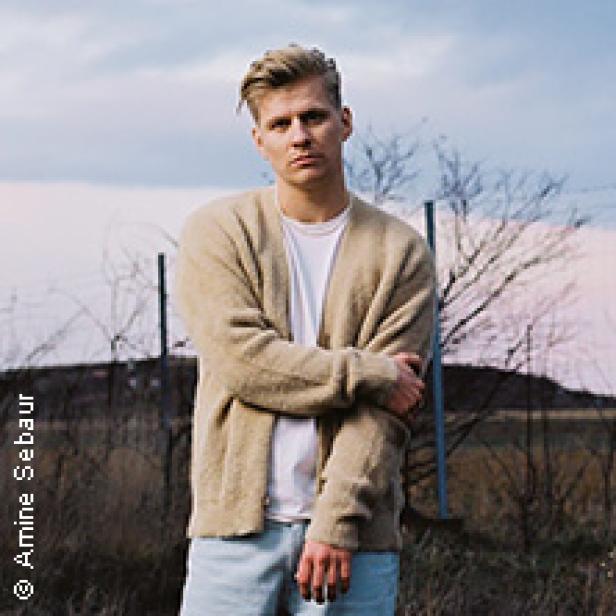 Ein Mann mit blonden Haaren und beiger Strickjacke steht vor einer Landschaft.