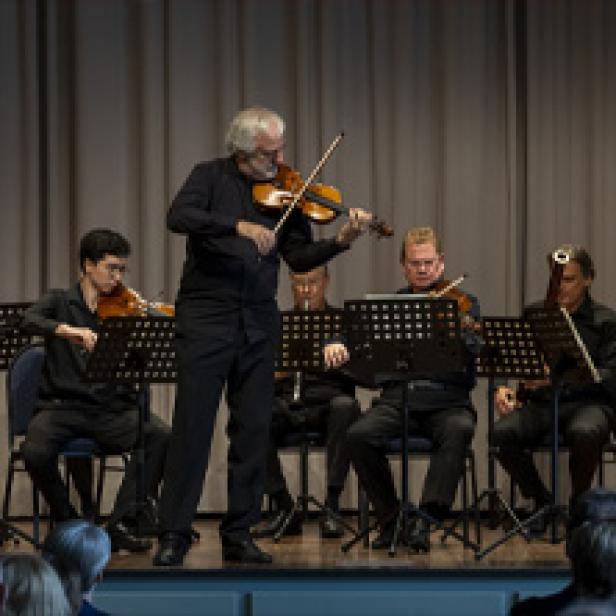 Ein Mann mit grauen Haaren spielt Violine vor einem Orchester.