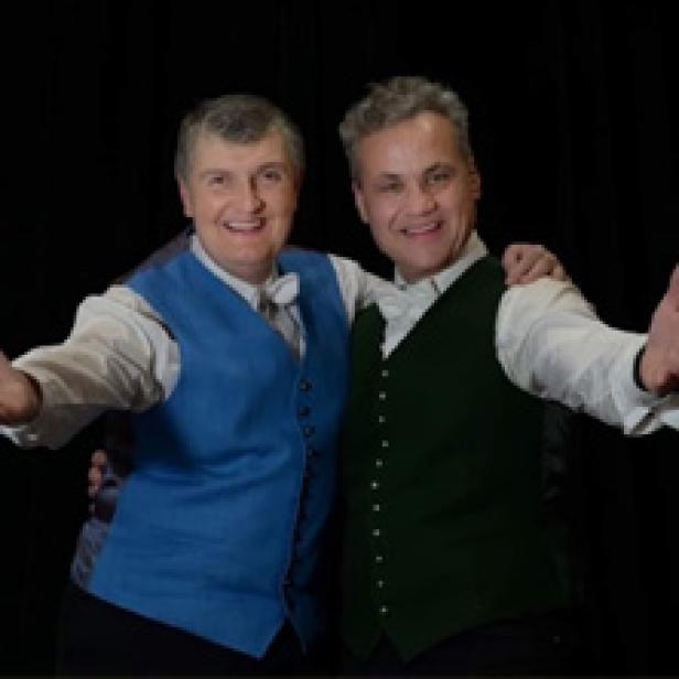 Zwei Männer in Westen lächeln und strecken die Arme aus.