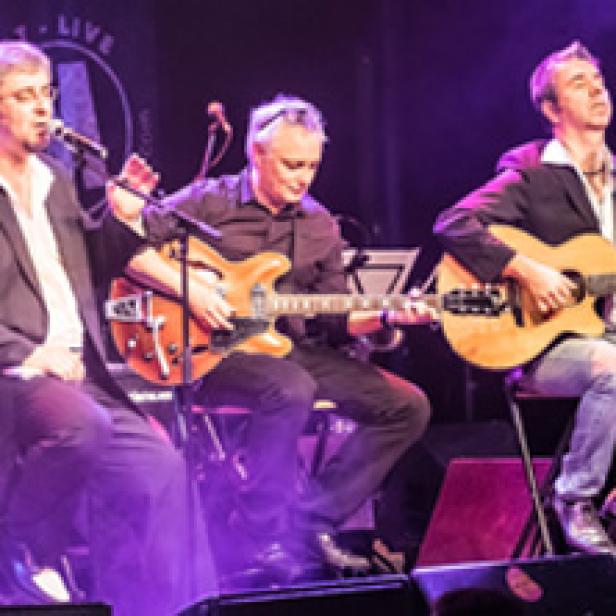 Drei Männer spielen Gitarre und singen auf einer Bühne mit violetter Beleuchtung.