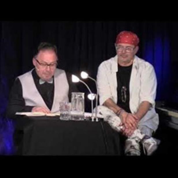 Zwei Männer sitzen an einem Tisch vor einem schwarzen Vorhang.