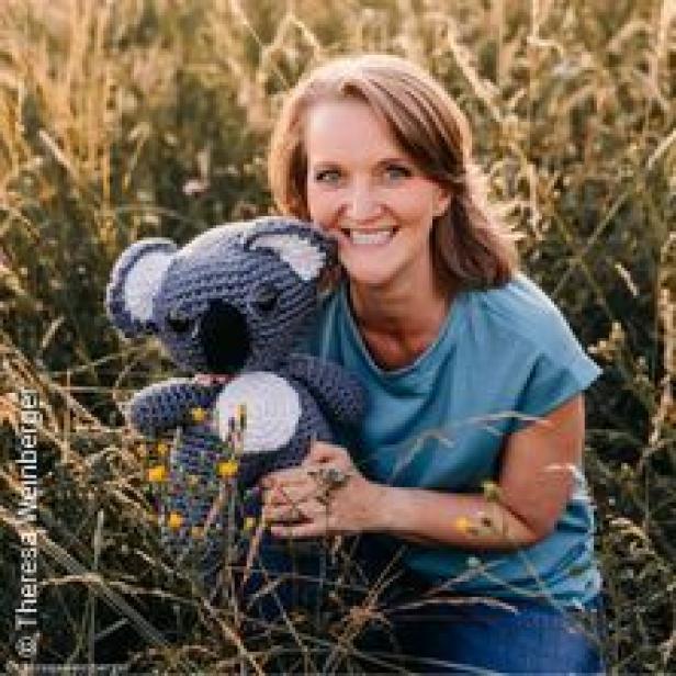 Eine Frau hockt in einem Feld und hält einen gehäkelten Koala-Bären.
