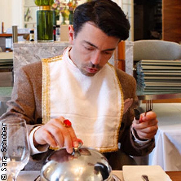 Ein Mann mit Lätzchen öffnet eine Speisehaube in einem Restaurant.