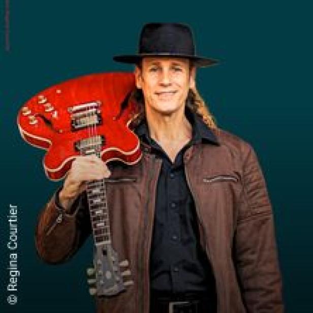 Ein Mann mit Hut und Gitarre posiert vor einem türkisfarbenen Hintergrund.