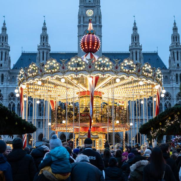 Ein beleuchtetes Karussell steht vor dem Wiener Rathaus, umgeben von Menschen und Marktständen.