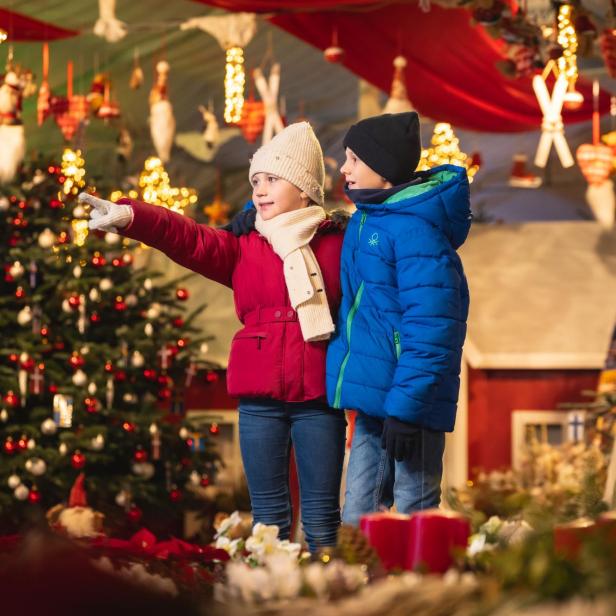 Zwei Kinder in Winterkleidung stehen vor einem Weihnachtsbaum in Hirschstetten und blicken auf einen Weihnachtsmarkt.